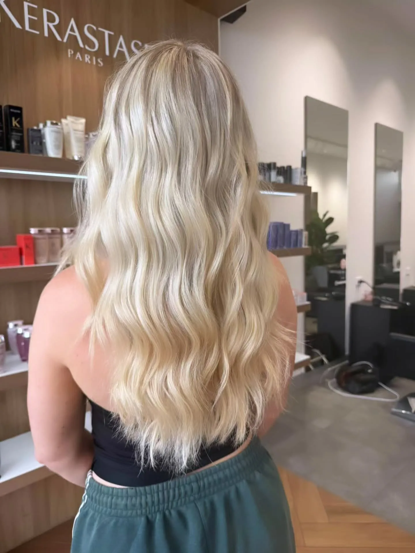 Back view of long beachy blonde waves in front of the Kérastase Paris wood-wall signage, mirror stations receding right