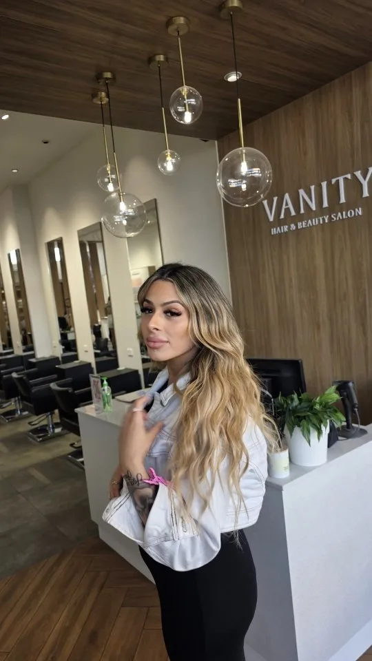 Woman posing under the Vanity wall sign with gold globe pendants and styling stations behind