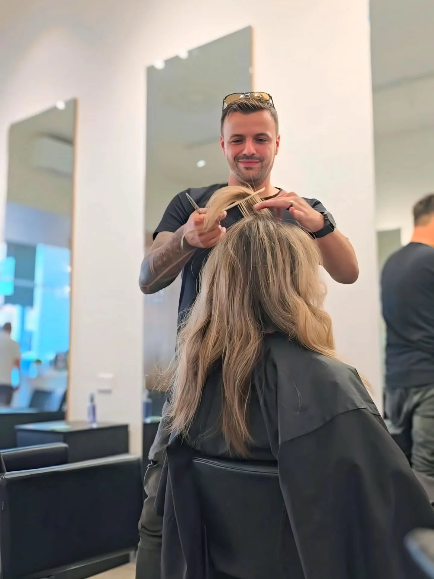 Male stylist mid-cut on a client's long blonde hair, bright mirrored salon behind