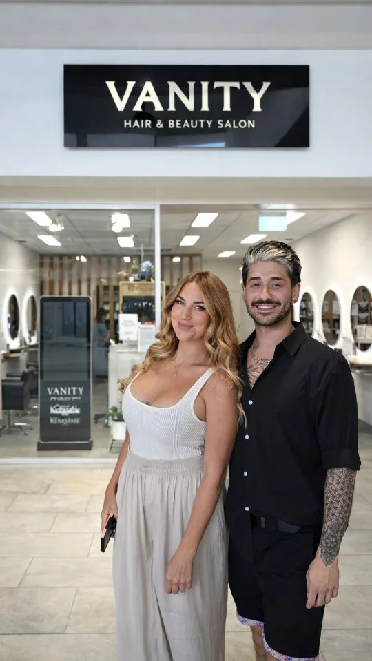A woman and a man standing at the Vanity salon entrance beneath the black fascia sign