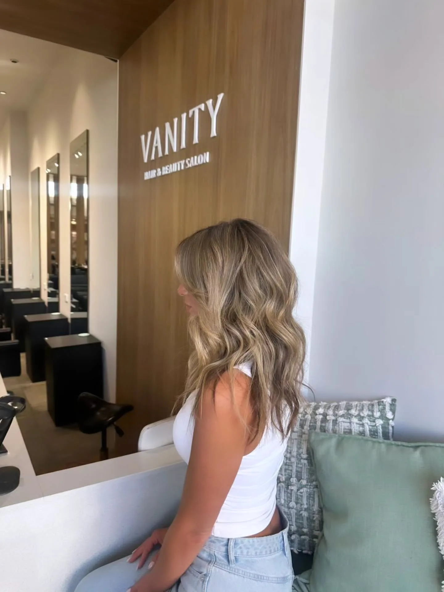 Woman seated beside the Vanity Hair & Beauty Salon wall sign, mirror stations receding behind
