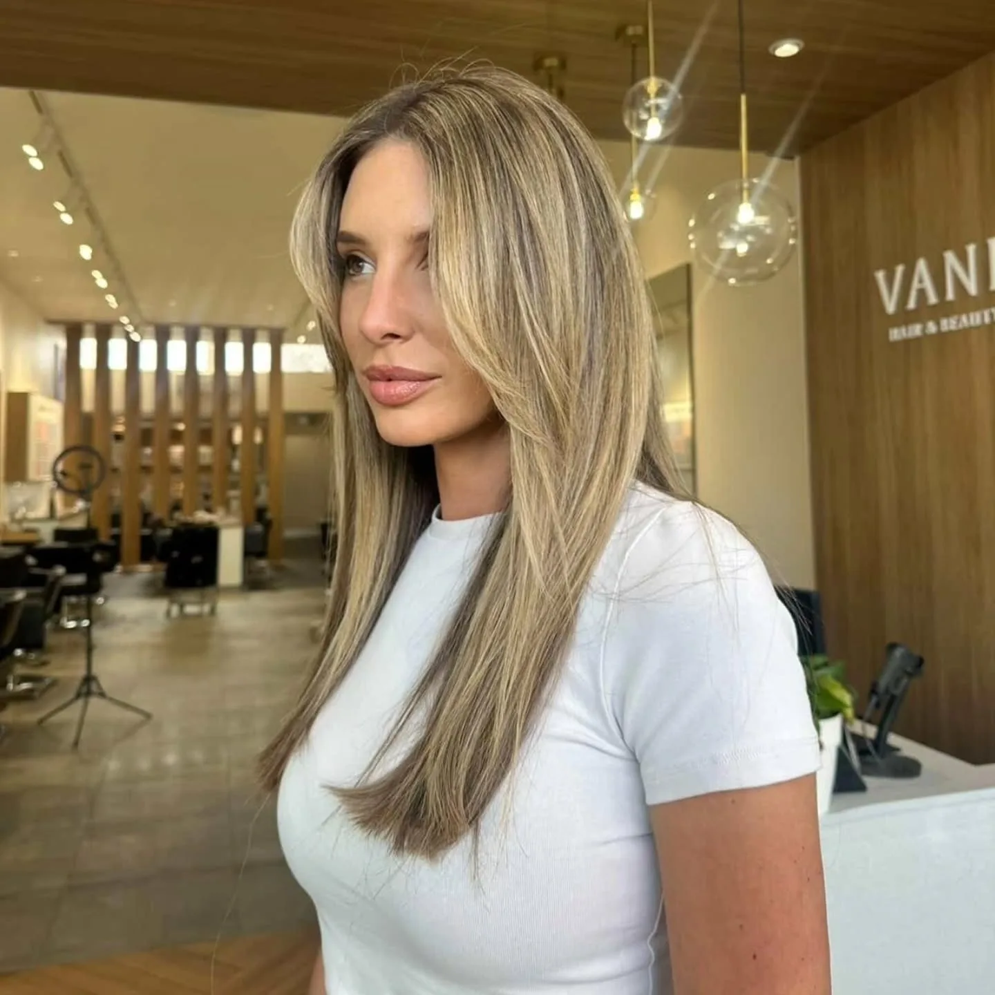 Woman with long blonde balayage beside the raised Vanity Hair & Beauty wall sign, gold globe pendants glowing behind